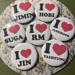 May include: A collection of seven white round buttons with the text "I ♥" followed by different names in black, and a red heart. The names include Jimin, Hobi, Suga, RM, Jungkook, Jin, and Taehyung.
