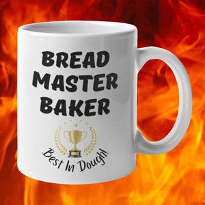 May include: White ceramic coffee mug with black text that reads "Bread Master Baker Best In Dough!" and a gold trophy with a laurel wreath.