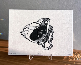 May include: Black and white linocut print of a cat inside a bag. The cat's face is visible, with two round eyes and whiskers. The print is on white paper, with the title "Bag" and the artist's signature.