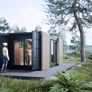 May include: A small, modern, black and wood-paneled house with a large window and a door open to reveal a person standing inside. The house is situated on a wooden deck and surrounded by green grass and trees.