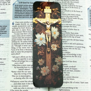 May include: A close-up of a religious bookmark featuring a crucifix with a white background and floral accents. The bookmark is made of wood and has a gold trim. The text on the bookmark reads "5:18 Good for Some" and "I have seen another evil".