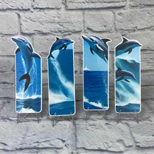May include: Set of four blue and black dolphin bookmarks with a blue ocean background. The dolphins are jumping out of the water. The bookmarks are on clear plastic stands.