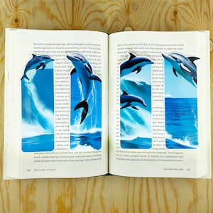 May include: An open book with a blue and white dolphin jumping out of the water on the left page and a blue and white dolphin jumping out of the water on the right page. The text on the left page reads "But until a couple decades ago, adult passengers could wantonly smoke cigarettes in that seat you're sitting in now. The nicotine headaches were real and pounding and the cabin was suffocating. ... everyone had different-colored hair and ate various things for lunch: sandwiches and burritos and corkscrew pasta. Nothing made me appreciate home more than leaving it. But I always felt..." The text on the right page reads "like a new person in the fall, as if I'd wrapped my mind a little further around the world over the summer in ways my classmates did see and couldn't understand. ... ing directions when you can't read the language. Traveling puts you in other people's shoes. It teaches you compassion and empathy and, greatest of all, humility. It's impossible to visit a..."