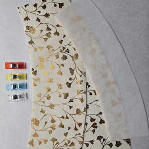 May include: Two pieces of fabric with a gold leaf pattern on a white background. The fabric is layered on top of a white piece of interfacing. There are four colorful binder clips on the left side of the image.