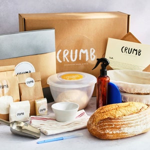 May include: A sourdough bread baking kit with a brown cardboard box, a white bowl with dough, a loaf of bread, a spray bottle, a proofing basket, a metal scoop, a kitchen towel, and a blue scraper. The box has the word "CRUMB" printed on it.