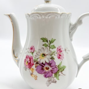 Puede incluir: Tetera de porcelana blanca con un diseño floral con flores rosas, moradas, blancas y amarillas. La tetera tiene un borde dorado alrededor de la tapa y el pico.