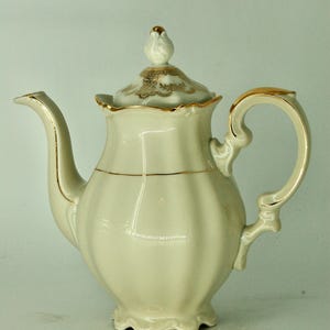Może przedstawiać: Kremowy porcelanowy imbryk ze złotymi detalami. Imbryk ma zakrzywioną wylewkę, pętlę uchwytu i dekoracyjną pokrywkę z kwiatowym guzikiem. Pokrywka i uchwyt są ozdobione złotem.