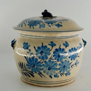 Könnte beinhalten: Ein beigefarbener Keramiktopf mit Deckel, verziert mit blauen Blumenmustern. Der Topf hat zwei Griffe und einen kleinen, dunkel gefärbten Knopf auf dem Deckel. Das Blumenmuster umfasst große und kleine Blüten.