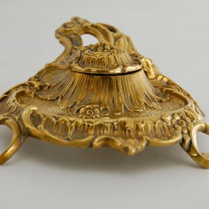 Könnte beinhalten: Ein goldfarbenes Tintenfass aus Metall mit einem verzierten Reliefmuster aus Blumen- und Schnörkelmotiven. Das Objekt verfügt über einen zentralen runden Deckel, einen breiten Boden mit drei geschwungenen Beinen und eine texturierte Oberfläche.