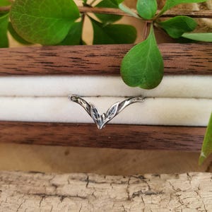 May include: A silver ring featuring a leaf design. The ring has a V-shape with three leaves at the centre. It is displayed in a wooden box with a white interior, with green leaves in the background.