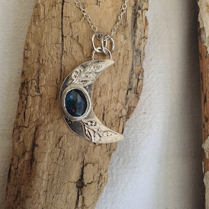 May include: A silver crescent moon pendant necklace featuring an oval faceted dark blue gemstone set in the centre. The metal surface of the moon is textured with engraved leaf patterns and is suspended from a delicate silver chain link.