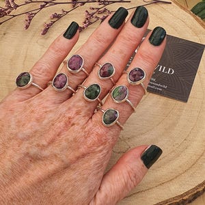 May include: A hand displaying multiple silver rings, each featuring oval gemstones in red and green hues. The rings are presented on a wooden surface. A business card with the word "Wild" is visible. The nails are painted dark green.