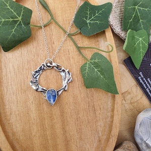 May include: A silver pendant necklace with a textured, open circular design and a blue, teardrop-shaped gemstone. The necklace is suspended from a fine chain. The setting is a wooden surface with green leaves.