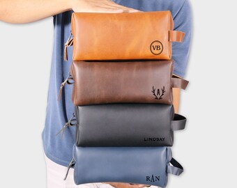 May include: Four leather toiletry bags stacked on top of each other. The top bag is brown with the initials "VB" in a circle. The second bag is dark brown with the initial "A" and antlers. The third bag is black with the name "LINDSAY". The bottom bag is blue with the initials "RAN".