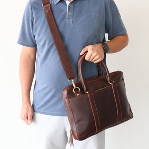 May include: A brown leather briefcase with a long adjustable strap. The briefcase has a zippered closure and a flap with a buckle.