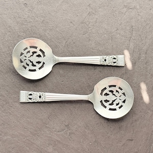 Puede incluir: Dos cucharas de servir plateadas con diseños florales calados ornamentados en el cuenco. Los mangos tienen un diseño acanalado con extremos decorativos. Las cucharas se muestran sobre una superficie gris oscuro.