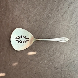 May include: A silver-colored serving spoon with a slotted, oval-shaped head featuring a sunburst design. The handle has an ornate, decorative pattern. The spoon is set against a dark gray, textured surface.