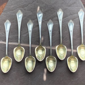 Puede incluir: Ocho cucharaditas de plata con mangos adornados y cuencos chapados en oro. Las cucharas están dispuestas en fila sobre una superficie gris oscura.