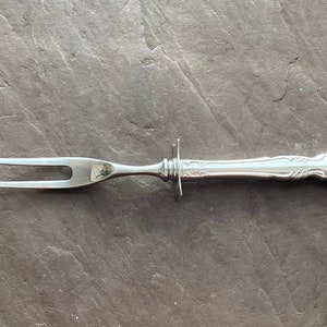 May include: A silver serving fork with a decorative handle. The fork has two prongs and a long, slender handle.