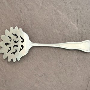 Könnte beinhalten: Silberfarbener Servierlöffel mit dekorativem, durchbrochenem Kopf. Der Kopf ist blütenförmig mit ausgeschnittenen Mustern. Der lange, schlanke Griff hat am Ende ein Blumendesign. Vintage Küchenutensil.