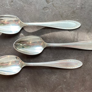 May include: Three silver spoons with ornate handles. The spoons are arranged in a row on a dark gray surface.
