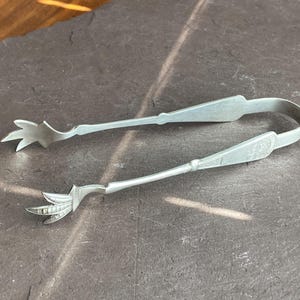 Puede incluir: Pinzas de azúcar de plata con un diseño floral decorativo en los extremos. Las pinzas están abiertas y listas para coger un terrón de azúcar.