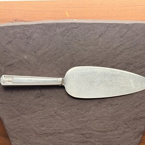 May include: Silver cake server with a decorative handle. The server has a wide, rounded blade and a detailed handle design. The server is placed on a dark gray surface.