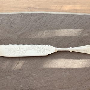 May include: A silver fish serving knife with a serrated edge and ornate floral engravings. The handle has a decorative design. The knife is lying on a dark gray stone surface.