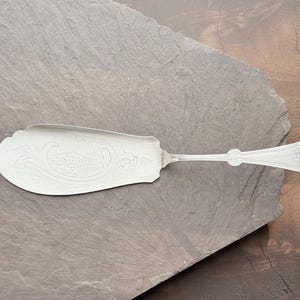May include: Silver-colored serving utensil with an ornate, fish-shaped design on the blade. The handle has a decorative pattern and a small, round detail. The utensil is placed on a dark gray stone surface.