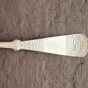 Puede incluir: Primer plano de un cuchillo de mantequilla plateado con grabados florales y geométricos ornamentados. El cuchillo tiene una hoja ancha y redondeada y un mango detallado. El diseño sugiere un estilo vintage o antiguo.