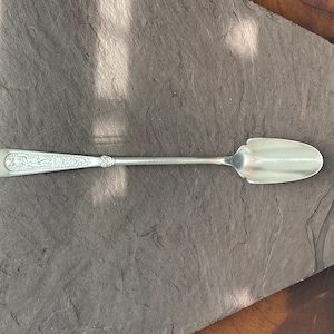 以下が含まれることがあります： 装飾的なハンドルと長く狭いスプーンを持つ銀製のサービングスプーン。スプーンは灰色のスレートの表面に置かれています。