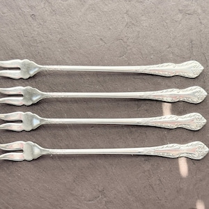 Peut inclure: Quatre fourchettes de service argentées à trois dents, disposées horizontalement. Les manches présentent un motif floral décoratif à l'extrémité. Les fourchettes sont présentées sur une surface gris foncé.