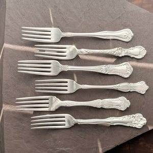 Peut inclure: Six fourchettes argentées aux manches ornés et détaillés sont disposées sur une surface gris foncé. Les fourchettes présentent des dents et des motifs décoratifs, suggérant un style vintage ou antique. L'arrangement est soigné.