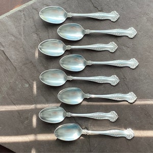 May include: Eight silver teaspoons with ornate handles arranged in a row on a dark grey slate surface.