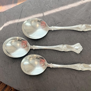 Puede incluir: Tres cucharas soperas plateadas con mangos ornamentados se disponen sobre una superficie gris oscuro. Las cucharas tienen un cuenco redondeado y detallados diseños florales en los mangos. Las cucharas reflejan el entorno.