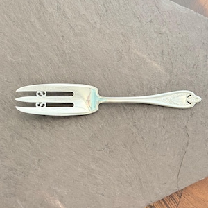 Puede incluir: Tenedor de servir plateado con tres púas y cortes decorativos. El mango tiene un diseño ornamentado en el extremo. El tenedor está colocado sobre una superficie de piedra gris.