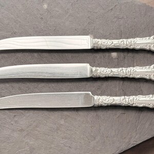 May include: Three silver butter knives with ornate, detailed handles. The blades are curved and smooth, reflecting the light. The knives are arranged on a gray surface, showcasing their elegant design.