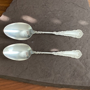 May include: Two silver spoons with ornate handles, lying on a dark gray textured surface.