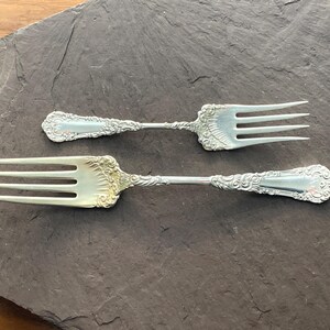 May include: Two silver forks with ornate handles, one with a gold finish, on a dark gray slate background.