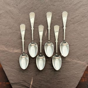 Puede incluir: Siete cucharas de plata dispuestas sobre una superficie gris oscuro. Cada cuchara tiene un mango detallado y un cuenco ovalado pulido. Las cucharas son probablemente para servir o comer, y son un artículo de mesa clásico.