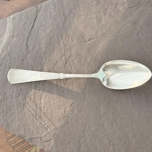May include: A silver spoon with an ornate handle and a large, oval bowl. The handle features engraved floral designs. The spoon is set against a dark gray stone surface, creating a contrast of textures and tones.