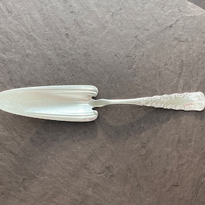 Puede incluir: Un servidor de pastel de plata con un mango adornado. El servidor tiene una hoja ancha y plana y una punta puntiaguda.