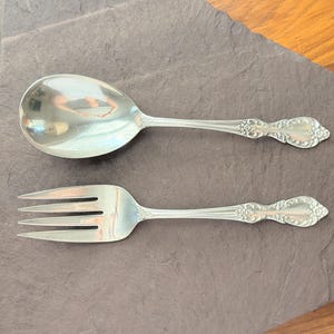 Op de afbeelding: Een zilveren opscheplepel en vork set met sierlijke bloemendetails op de handvatten. De lepel heeft een afgeronde kom en de vork heeft drie tanden. De set wordt gepresenteerd op een donkergrijze stenen ondergrond.