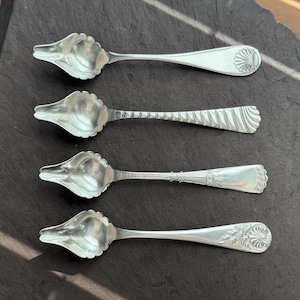 Könnte beinhalten: Vier silberne Löffel mit muschelförmigen Schalen und dekorativen Griffen. Jeder Löffel hat ein einzigartiges Griffdesign, einschließlich floraler und geometrischer Muster. Die Löffel sind auf einer dunklen, strukturierten Oberfläche angeordnet.