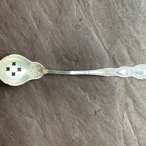Puede incluir: Una cuchara de azúcar de plata con un mango decorativo y un cuenco perforado con cuatro agujeros. La cuchara está diseñada para recoger terrones de azúcar.