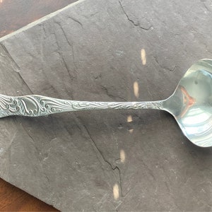 Könnte beinhalten: Eine silberne Schöpfkelle mit einem verzierten, gravierten Griff. Die Kelle hat eine runde Schale und einen langen, schlanken Griff.