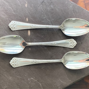 May include: Three silver spoons with ornate handles and a polished finish. The spoons are arranged in a row on a dark gray surface.