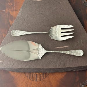 Puede incluir: Tenedor y servidor de pastel plateados con detalles florales ornamentados. El tenedor tiene tres púas y el servidor tiene una hoja ancha y plana. Ambas piezas se exhiben sobre una superficie triangular gris oscuro.