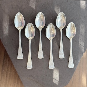 May include: Six silver spoons arranged on a dark gray surface. The spoons have a reflective, polished finish, with a textured pattern on the handles. The spoons are likely for serving or eating.