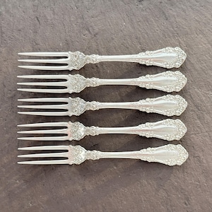 Può includere: Cinque forchette d'argento con dettagli floreali ornati. Le forchette hanno tre rebbi e un manico decorativo. Le forchette sono disposte in fila su una superficie scura e strutturata. Le posate sono probabilmente per un ambiente formale.